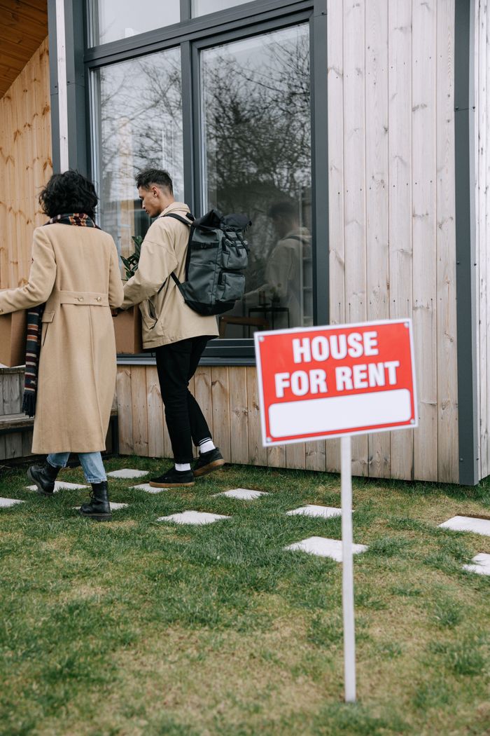 A couple enters a modern house for rent with a prominent sign on a grassy lawn.